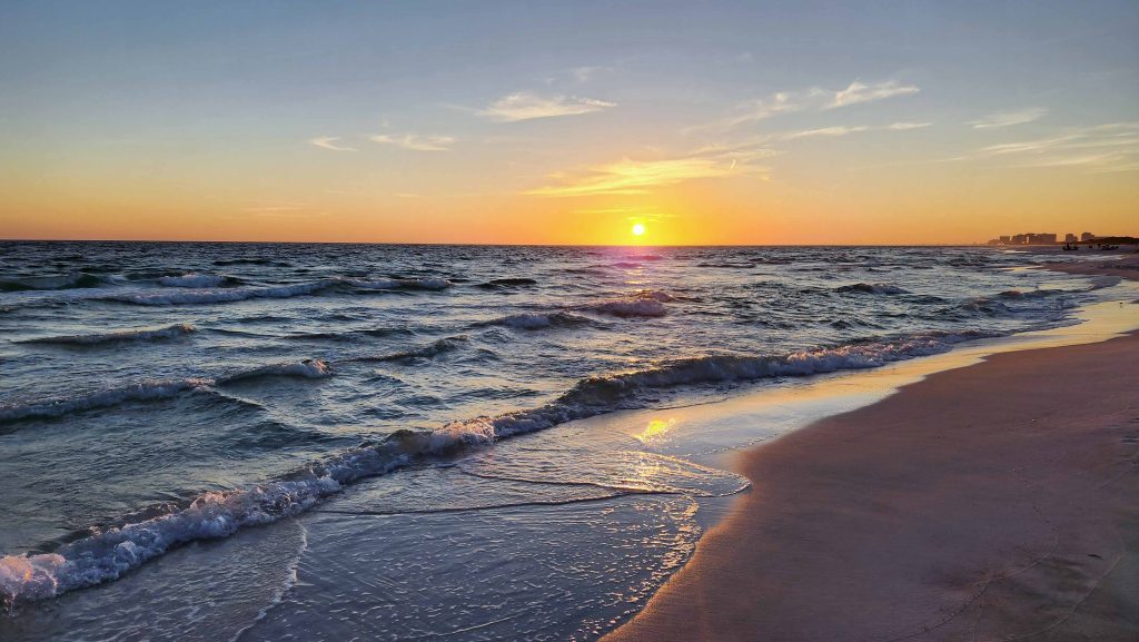 White sandy beach with waves splashing while sun sets in distance.