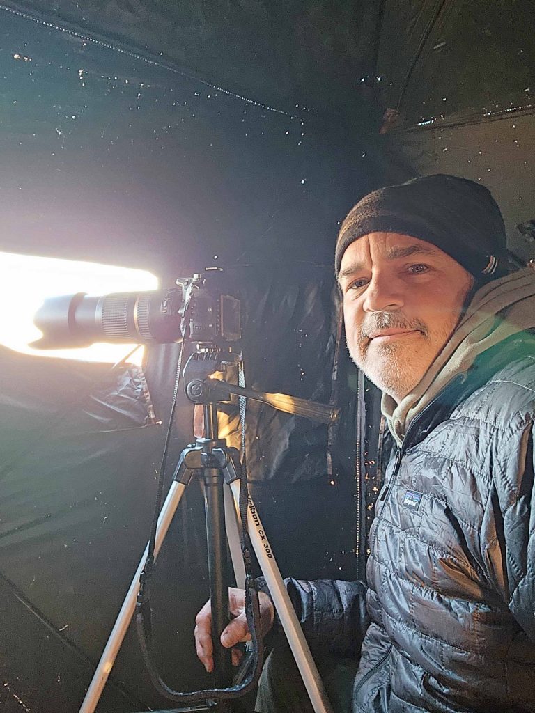 Brad Saum bundled for cold weather sitting by a camera on a tripod in a bird blind.