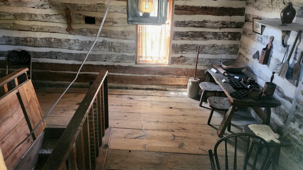 Inside view of pre-civil war era log cabin with furniture and a stairway leading from the floor below ground to a tunnel.