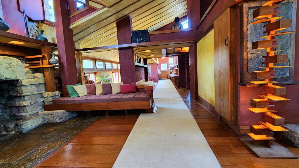 Open floorplan in working area of Taliesen showing edge of stone fireplace on one side and a unique wood lamp on right side with a sofa in the middle and views into additional rooms.