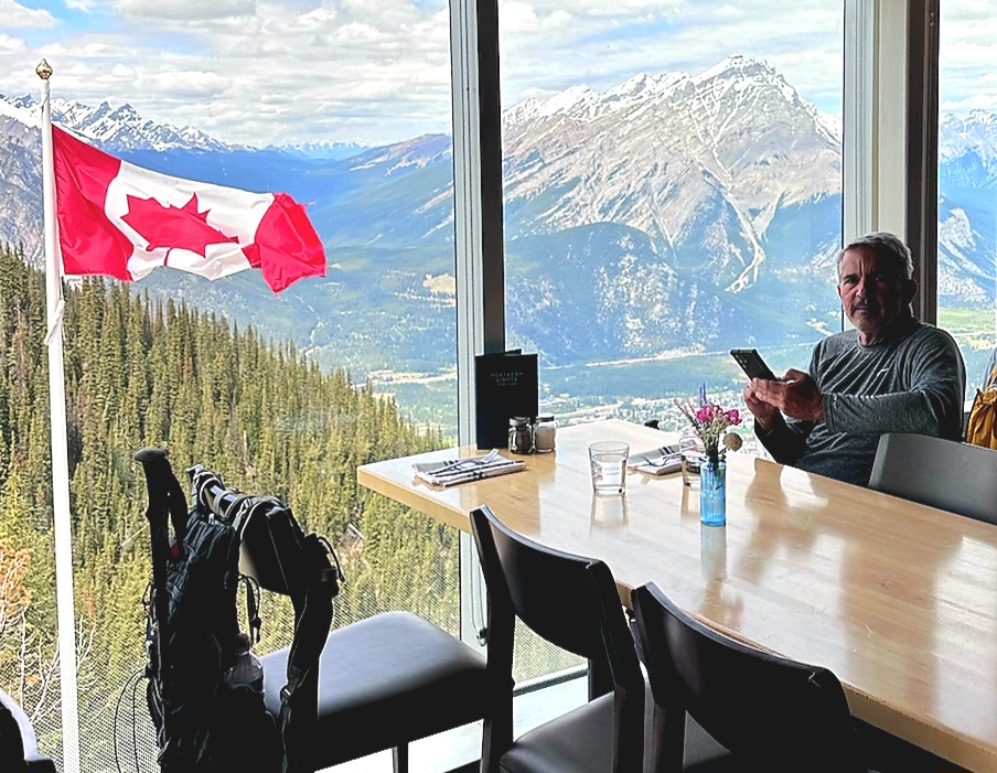 Brad Saum sitting at table at Northern Lights restaurant with mountains in the background and a Canadian flag flying